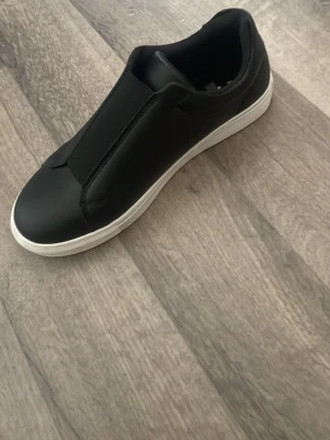 Svarta finskor med vit sula - Stilrena svarta slip-on sneakers med slät ovandel och vit gummisula. Skon har minimalistisk design utan snörning och är tillverkad i ett material som ser ut som syntetläder. Perfekt för dig som gillar en clean och modern look. Använda 1 gång.