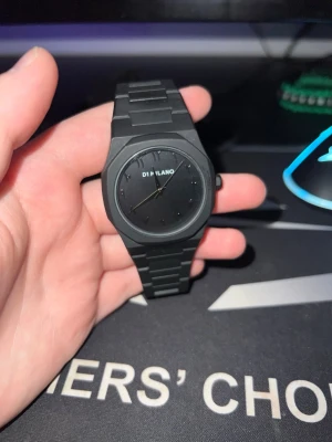 Svart klocka från D1 Milano - Stilren svart klocka från D1 Milano med mattsvart urtavla och matchande länkarmband. Klockan har minimalistisk design utan siffror och en diskret logga på urtavlan. Perfekt accessoar för dig som gillar en clean look.