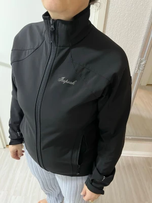 Svart vindjacka från Icepeak - Snygg svart vindjacka från Icepeak med hög krage och dragkedja framtill. Jackan har två sidofickor med dragkedja och diskret logga på bröstet samt en patch på ärmen. Tillverkad i ett slätt, vindtätt material som passar perfekt för blåsiga dagar.