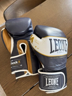 Leone boxningshandskar 14 oz - Snygga boxningshandskar från Leone i blått, vitt och brunt läder. Handskarna har tydlig Leone-logga, kardborrestängning och är märkta med 14 oz. Perfekta för träning eller sparring och har en limiterad Shock X-design. Använda ca 12-15 gånger.
