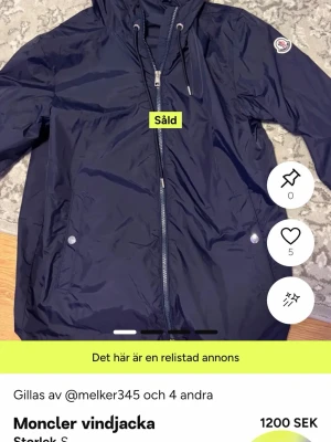 Mörkblå vindjacka från Moncler - Snygg mörkblå vindjacka från Moncler med dragkedja framtill, två sidofickor med tryckknappar och justerbar huva. Jackan har Moncler-logga på ärmen och är tillverkad i ett lätt, vattenavvisande material. Perfekt för blåsiga dagar.