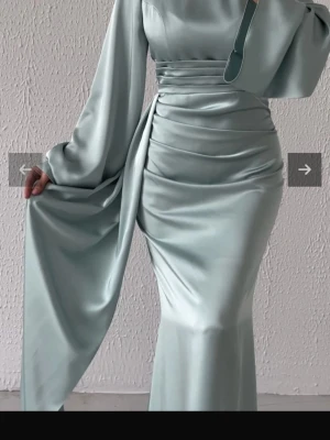 Ljusgrön långklänning i satin - Elegant ljusgrön långklänning i glansigt satinmaterial med markerad midja och draperade detaljer. Klänningen har långa vida ärmar och en mjuk, flytande siluett som ger en lyxig känsla. Perfekt för dig som vill sticka ut med stilren elegans.