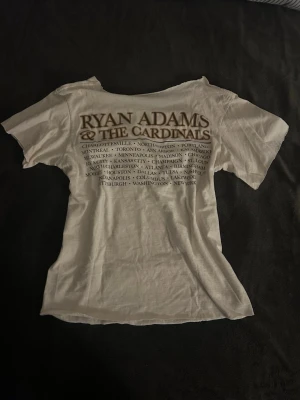 Ryan Adams & The Cardinals t-shirt - Vit t-shirt med tryck från Ryan Adams & The Cardinals. Framsidan har ett stort motiv av en ros i brunt och baksidan listar turnéstäder. Klassisk passform med rund hals och korta ärmar. Perfekt för dig som gillar bandmerch och vill sticka ut.