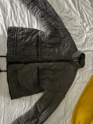 Zara jacka  - Snygg olivgrön/grå vindjacka med quiltat mönster och dragkedja framtill. Jackan har två stora fickor och elastisk nederkant. Materialet är tunt och lätt, perfekt för lager-på-lager. Enkel och stilren design som passar till det mesta.