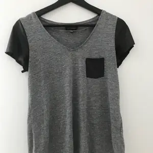 Grå t-shirt med svarta ärmar i tunnt skinnliknande material i storlek 36/38. Har använts en hel del och är lite sliten i ärmarna men annars är tröjan i bra skick.