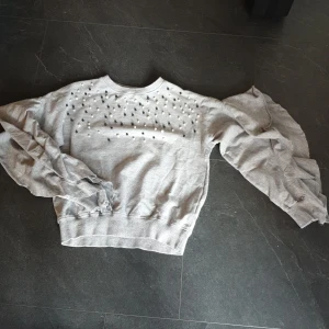 Tröja med pärlor - Grå sweatshirt med svarta och vita pärlor och volanger på ärmarna. Köpt i USA motsvarar storlek ca 152 cl.