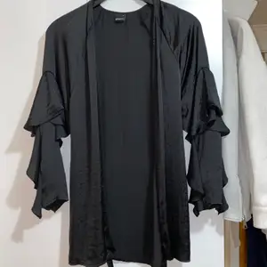 En svart kimono i satin från Gina Tricot. Den har volanger på ärmarna samt ett knytbälte i midjan. Funkar som ett nattplagg såväl som en kimono över en tröja till ett par byxor. Passar även xs.