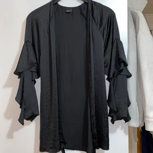 Satinkimono - En svart kimono i satin från Gina Tricot. Den har volanger på ärmarna samt ett knytbälte i midjan. Funkar som ett nattplagg såväl som en kimono över en tröja till ett par byxor. Passar även xs.