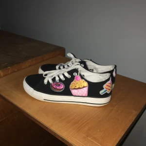 Svarta skor med gulliga märken - Ett par svarta Tamaris-skor i väldigt bra skick med 3 stickers i former av en doughnut en cupcake och en glass. Storlek 38 men jag skulle säga att de snarare är 37, möjligtvis 36. Köpare står för frakt som kostar 44kr.    