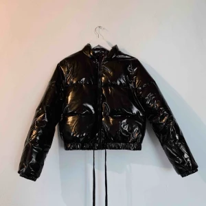  - Svart, glansig pufferjacka med dragkedja.