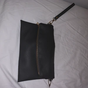Grå clatch - En grå väska som egentligen ska ha ett längre band men detta har tyvärr gått sönder, finns med ett mindre band som syns på bilden vilket gör att man ändå kan använda den. Långa bandet följer även med om man skulle vilja det.