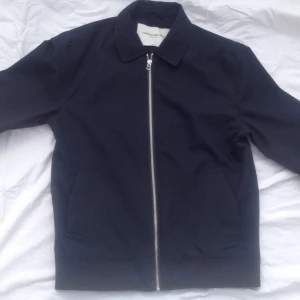  - Vårjacka i klassisk harringtonstil med vanlig krage. Felköp från årets vårkollektion och kan ses på märkets hemsida. Den heter Nathan jacket och kostar nu ca 1500 i butik. Mörkblå med dragkedja och muddar i ärmslut och midja. Fickor i sidorna samt innerfickor. Lätt nylonmaterial. Normal i storleken. Nyskick.