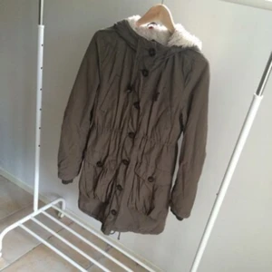  - Perfekt höstjacka i grön/brun färg från H&M divided. Luva med snöre som gör att man kan dra åt den. Muddar i armsluten. Innertyget på övre halvan samt inuti luvan i vitt Teddy tyg. I midjan finns ett gömt snöre som kan dras åt för att ge jackan önskad passform. Nederst på jackan finns även där ett snöre som kan dras åt. Längst ner på baksidan delas jackan lite för att inte bli för tight när man rör sig. 
Jackan knäpps med både dragkedja samt med knappar som knäpps över dragkedjan så den inte syns. 
Framtill två stora fickor som knäppa med knappar. 
Två mindre bröstfickor högre upp. 
Väl använd men fortfarande i bra skick. 

Frakt betalas av köparen. 