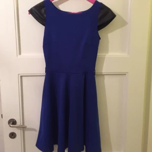  - Kornblå klänning med högmidja och knäppning bak i nacken. Ärmarna är gjorda i svart skinnimitation.