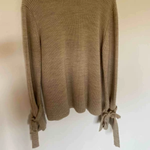  - Stickad tröja med rosetter runt handlederna. Färgen är lite missvisande på bilden. Tröjan är ljusbeige. Storlek L. Skickas spårbart och fraktkostnad på 63kr tillkommer 💝
