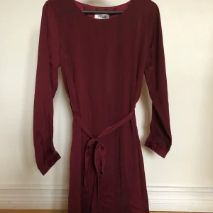 Vinröd klänning  - Vinröd klänning från märket DRY LAKE i siden eller silke med band runt midjan. Köpt för något år sedan, aldrig använd. Klänningen sitter ganska löst men får en snygg passform med bandet i midjan. Orginalpris 600 kr