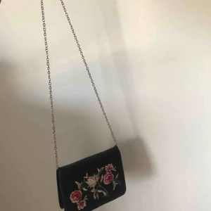  - Svart väska med blommor på framsidan, tunn kedja som väskband