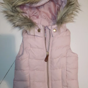 Lyxig täckväst i nyskick strl 92 - Knappt använt denna goding! Puderrosa och pälsen runt huvan är i jättefint skick.  Fluffig så den är säkert rätt varm, kanske lagom för denna milda vintern och en sväng på stan!  Djur och rökfritt hem. Samfraktar gärna,kika på mina övriga annonser!