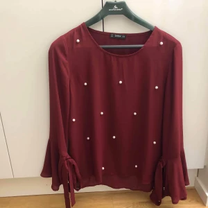 Topp med pärlor  - En vinröd topp med vita pärlor på framsidan samt snygga knytdetaljer längs ärmarna. Har aldrig använts! Säljes då jag är i behov av pengarna. 