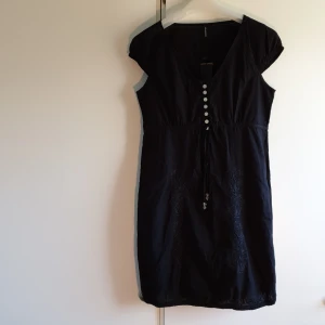 Ny Klänning  - Svart klänning med broderat blommönster fram. Band att knyta in i midja med pärlor. 