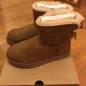 UGG Mini 39 - Säljer mina fina Ugg modell mini Bailey Bow II i klassiska färgen chestnut. Använda enstaka gånger men tyvärr för stora för mig! I nyskick.