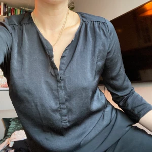 Svart blus i glansigt material - Perfekt till kontoret eller till vardags! Glansig svart blus med knappar fram. Bra skick! Frakt tillkommer