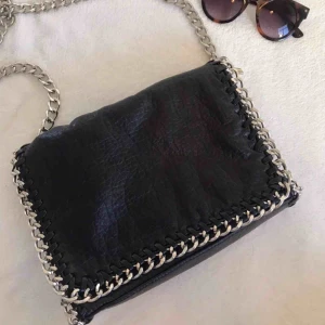  - Ursprungspris: 500kr Trendig Tiamo Pricilla väska, ”svart axelremsväska i skinnimitation kantad med silverfärgad kedja. Axellänk 105 cm. Längd 28 cm, höjd 18 cm, bredd 5 cm.” (Scorett). Ett fack med dragkedja på insidan o utsidan 