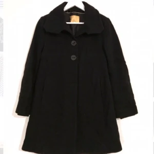  - Svart kappa i 70% ull med mjukt foder inuti! Två stora knappar längst upp, och dold knäppning nedåt. Muddar längst ned inuti ärmarna. Litet dammig då den legat med annat ylle. Annars i fint skick!  Kan mötas i Stockholm eller skicka mot fraktkostnad! ✨🌸✨