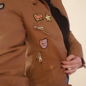 Skinnjacka stl m/l - Thriftad skinnjacka (fake ofc) i ljusbrunt med påsydda märken. Lite lång i ärmarna för mig som är 170 cm men passar perfekt över axlarna. Superfint skick!