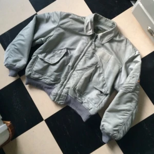  - ALPHA INDUSTRIES B-15 fint Silvergrå bomberjacka med krage. Helt ok skick. Inga fläckar eller hål/skador. Som alla pilotjackor är den bred men kort. Jag har normalt storlek L  men den är helt ok på mig om en vill vara lite baggy.