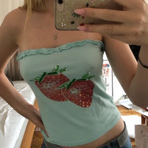 Ljusblå topp med jordgubbar - Så söt topp utan axelband med jordgubbar och paljetter. Köpt på plick men är tyvärr lite stor på mig som har s så den kommer inte till användning. Kan mötas i Stockholm annars betalar köpare frakt. 