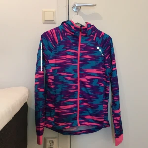 Tråningsjacka från soc - En tun träningsjacka från märket soc, köpt på stadium. Dragkedja längs hela framsidan, avtagbar luva och hål för tummarna längst ner på armen. Färgerna skiftar i rosa, lila och blått. Lite regntåligt material. Storlek 158-164. Frakt tillkommer
