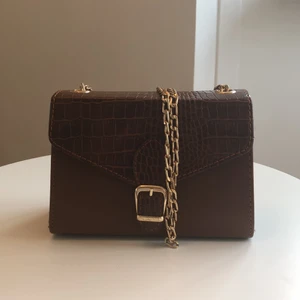 Snygg brun axelväska med lång guldkedja från BOOHOO - Väskan har stort utrymme och man kan ha mycket i den. Den hänger inte överdrivet långt ner. ”Snakeskin” på övre delen av väskan och även på sidorna. Den har lite avsaknad av guldfärg på spännet men inget som syns överdrivet mycket. Frakt ingår ej i pris