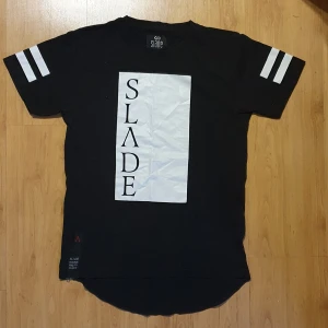 Snygg Slade t-shirt - Slade t-shirt med vitt tryck på framsidan och på armarna samt dekorativ dragkedja på sidan. 100% bomull. Alldrig använd. 350 kr ny säljer för 170 kr.