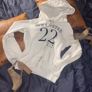 Hollister huvtröja med blixtlås  - Material: bomull till mestadels, polyester och foder. Färg: vit med marinblå brodering. Huvtröja från hollister med lite sliten brodering på vänstra tvåan (vänster från att man har på sig den). Silvrig dragkedja, då hollisters alla koftor knäpps åt fel håll kan det vara svårt första gången men den är hel. 