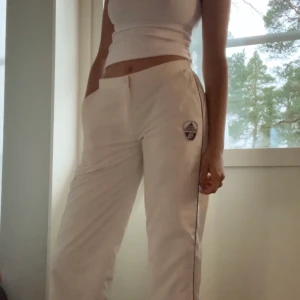 Vita Adidasbyxor  - Ett par skitballa vita lågmidjade Adidasbyxor. Köpta från humana. De har dragkedjor längst ner på vardera sida. Storlek 38 men passar nog 36. Går ner till anklarna, är 173 cm