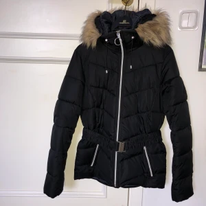 Vinterjacka - Varm vinterjacka med fakepäls. Skärp i midjan vilket gör jackan figursydd, avtagbart. Ganska lång i modellen. Mycket bra skick. Strl 36/38