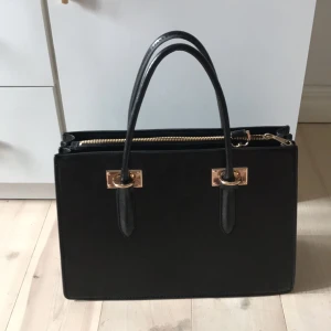  - Väska från h&m (nyskick!) Helt oanvänd, svart med gulddetaljer  Två handtag och lång axelrem som är avtagbar.  Dragkedja i mitten och två öppna fack med innerfack för mobil/ småsaker Svart innerfoder   25cm hög och 33 cm bred 