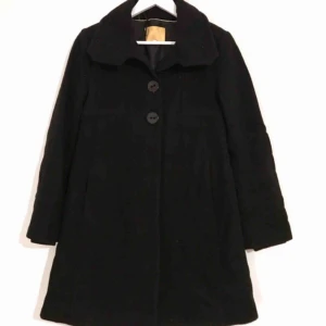  - Svart kappa i 70% ull med mjukt foder inuti! Två stora knappar längst upp, och dold knäppning nedåt. Muddar längst ned inuti ärmarna. Vårjacka. Litet dammig då den legat med annat ylle, och behöver strykas. Men annars i fint skick!