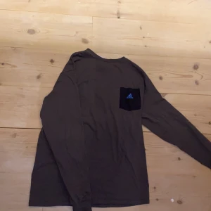 Långärmad t-shirt adidas mörkgrå, svart ficka och blå adidaslogga - långärmad mörkgrå adidas tröja med svart ficka och mellanblå adidaslogga på. 🖤 Mycket bra skick. Storlek XS men på mig som är en XS/S tycker jag att den sitter oversized och snyggt. Men är ganska smal längst ner i ärmarna. 100% cotton (bomull). Mate
