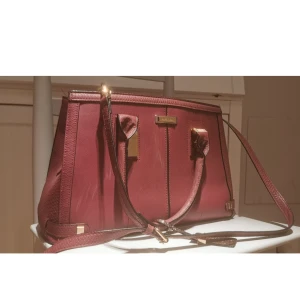 River Island vinröd väska  - En vackert vinröd väska från River Island, möjligt att både bära den i handen såsom ovanpå axeln. Köptes in tidigare under sommaren i år på marketplace men den passar inte riktigt till min stil! Rymlig och lätt sorteringsbar som är fördelade i tre i rad. Mitten facket är det enda förslutningsbara med draghedla. Kan anas se konturer av  krokodilhud i yttre materialet. Måtten: bredden är på längsta sidan 35 cm och kortaste på 13 cm, höjd en är 24 cm och djupet 15 cm. Bra skick och hållbar. 