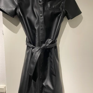 Klänning i skinnimitation - Så fin klänning i skinnimitation. Sitter ganska löst på kroppen och man får ett bälte till, väldigt bra skick. Frakt 50kr