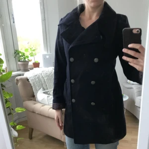  - Stilren kappa i ull med dubbelknäppning. Perfekta höst/vinterkappan. Färgen är mörkblå, nästan svart. Fantastiskt foder. Inköpt för några år sen på HM men har mestadels hängt i garderoben, använd kanske 5 gånger. Varm och skön, kragen går att ha upp eller nedvikt. Passar perfekt på en 36a, sitter tight på en 38a och lite loose på en 34a.   Köparen står för frakten på 70 kr.