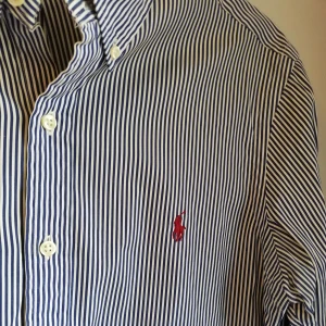 Herrskjorta från Ralph Lauren. - Herrskjorta från Ralph Lauren i storlek M; slim fit. Jättefint skick. Blå och vit-randig med ett vinrött märke. Material: 100% bomull. Frakt tillkommer. Bara att skriva om ni undrar något. 