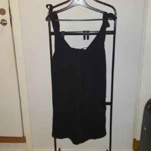  - Svart byxdress med knytband över axlarna och en öppning i ryggen och blixtlås bak. 