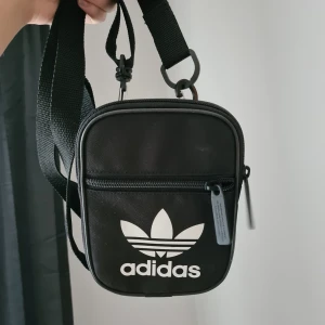 Adidas väska - Fin liten väska där man kan förvara mobilen, plånboken mm. Har två st fack. Knappt använd.                               Längd/höjd?: 15,5 cm. Bredd: 11,5 cm                                         Frakt tillkommer