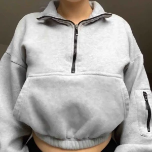  - Ljusgrå croppad hoodie. Lång justerbar dragledja längst bysten. Stor ficka vid magen samt på vänster arm. Kardborrespännen längst ner på armarna. Färgslitage vid dragkedjespännena, annars hur fin som helst! 