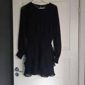 Klänning - Svart klänning i tunt tyg med glansiga prickar. Den har underklänning så bara ärmarna är lite genomskinliga. Smock i midjan och ärmsluten. Typ muddar.