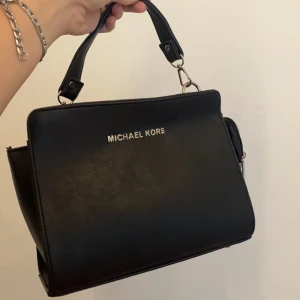 Handväska Michael Kors - Svart väska med silverdetaljer, väldigt stilren. Säljer pga inte min stil. Väskan är köpt för ganska länge sen så är inte 100% på om den är äkta, så säljer till ett ganska lågt pris för att inte lura någon. 
