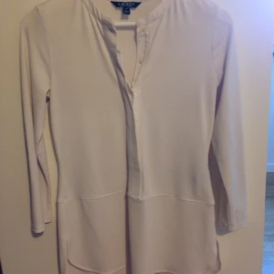  - Lauren by Ralph Lauren blus i vit trikå vilket gör den elastisk. Dold knappslå halv framtill och rundhalsad kraglös. Ärm 3/4 smal. Väldigt fin passform och i mycket gott skick. Tvättad 2 ggr och lite använd. Köpt på best of brands 2016. 